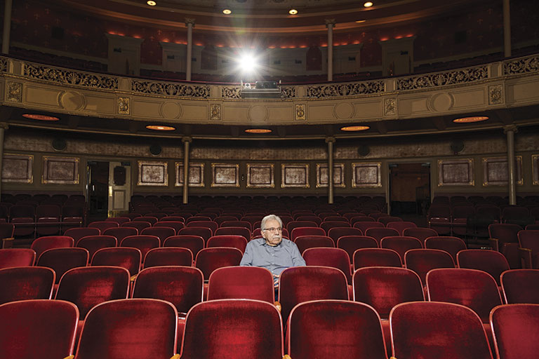 An older man sits alone among rows of red velvet seats in an ornate, warmly lit historic theater, creating a quiet, contemplative mood.