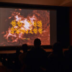 Audience silhouettes sit in a dark auditorium, watching a large screen display fiery red imagery with glowing yellow script, creating a dramatic, mysterious mood.