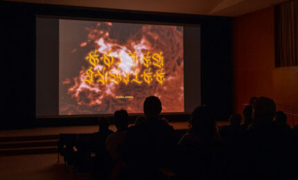 Audience silhouettes sit in a dark auditorium, watching a large screen display fiery red imagery with glowing yellow script, creating a dramatic, mysterious mood.