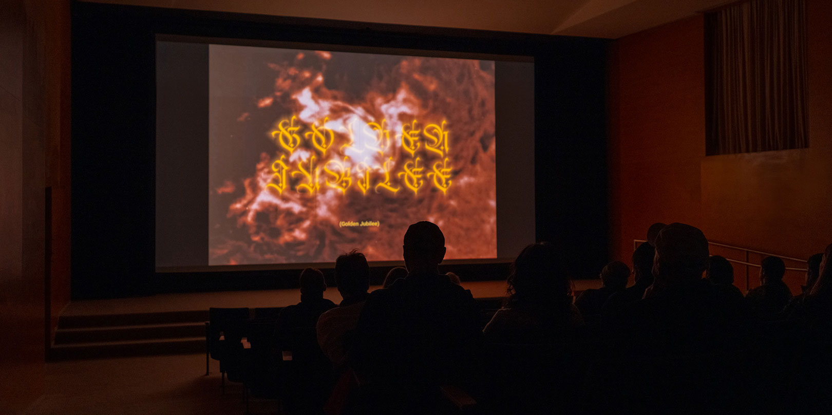 Audience silhouettes sit in a dark auditorium, watching a large screen display fiery red imagery with glowing yellow script, creating a dramatic, mysterious mood.
