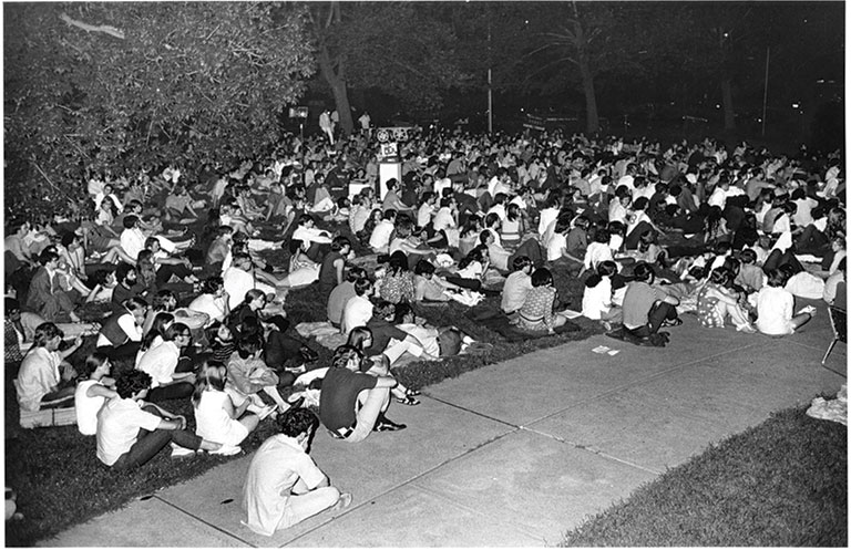Large nighttime crowd seated on grass and a sidewalk, many holding signs; the peaceful gathering conveys solidarity, reflection, and concern.