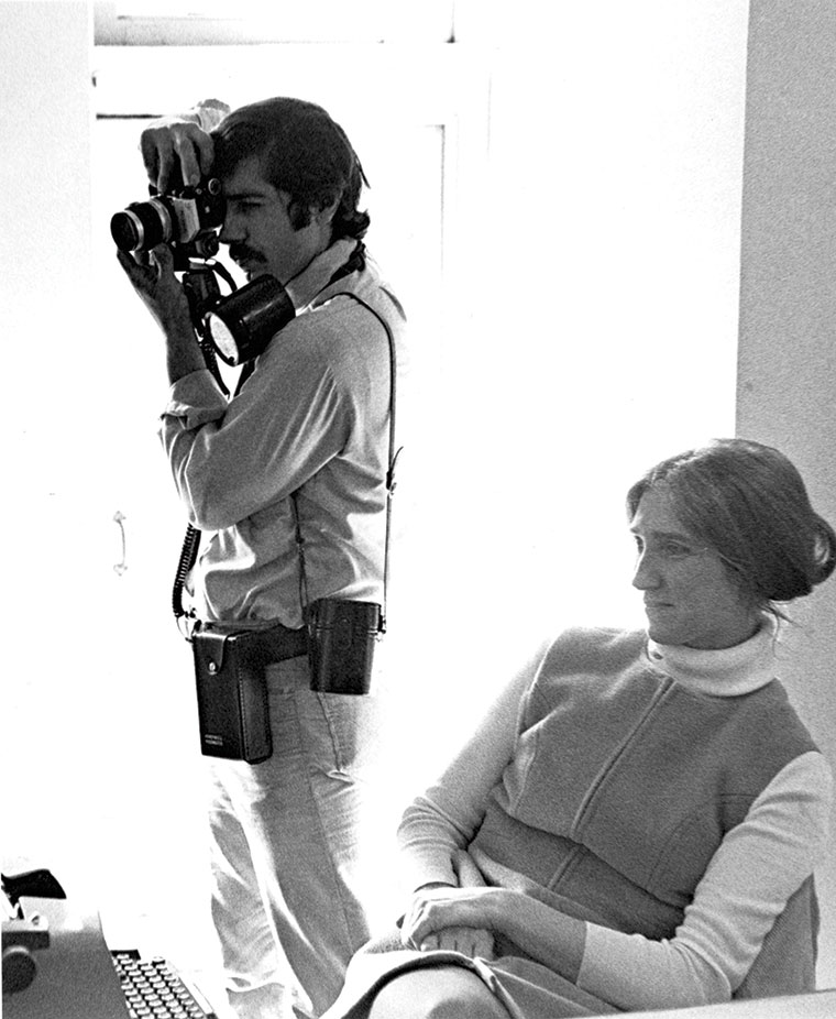 Black-and-white photo of a focused photographer using a shoulder-mounted camera while a seated woman watches calmly in a bright room.