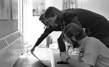 Two people crouch in a hallway as a woman uses a cane to point at the floor, carefully inspecting the space in a serious black-and-white scene.