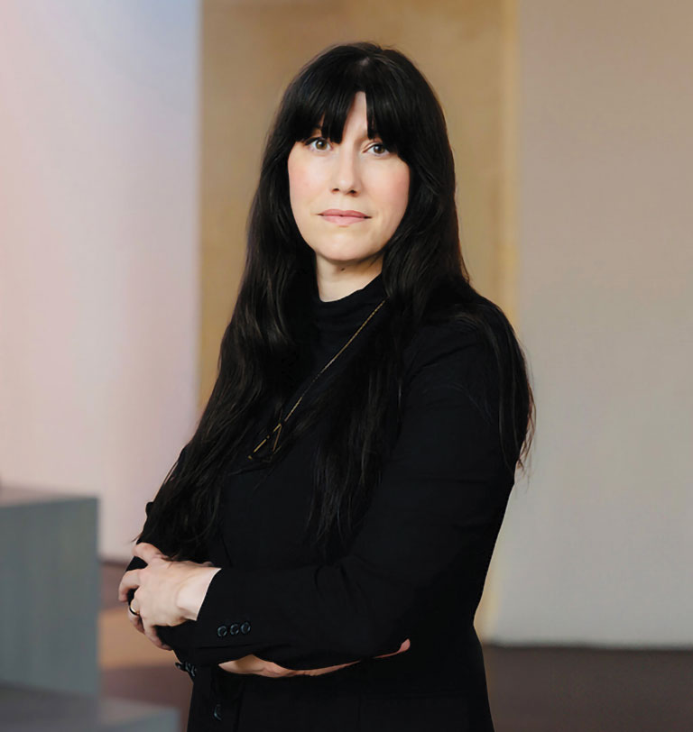 Portrait of a woman with long black hair in a black outfit, standing with arms crossed indoors; her calm expression creates a poised tone.