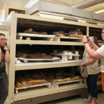 Visitors examine shelves of breads and pastries in a bakery display rack, while a woman photographs the baked goods in a busy, curious scene.