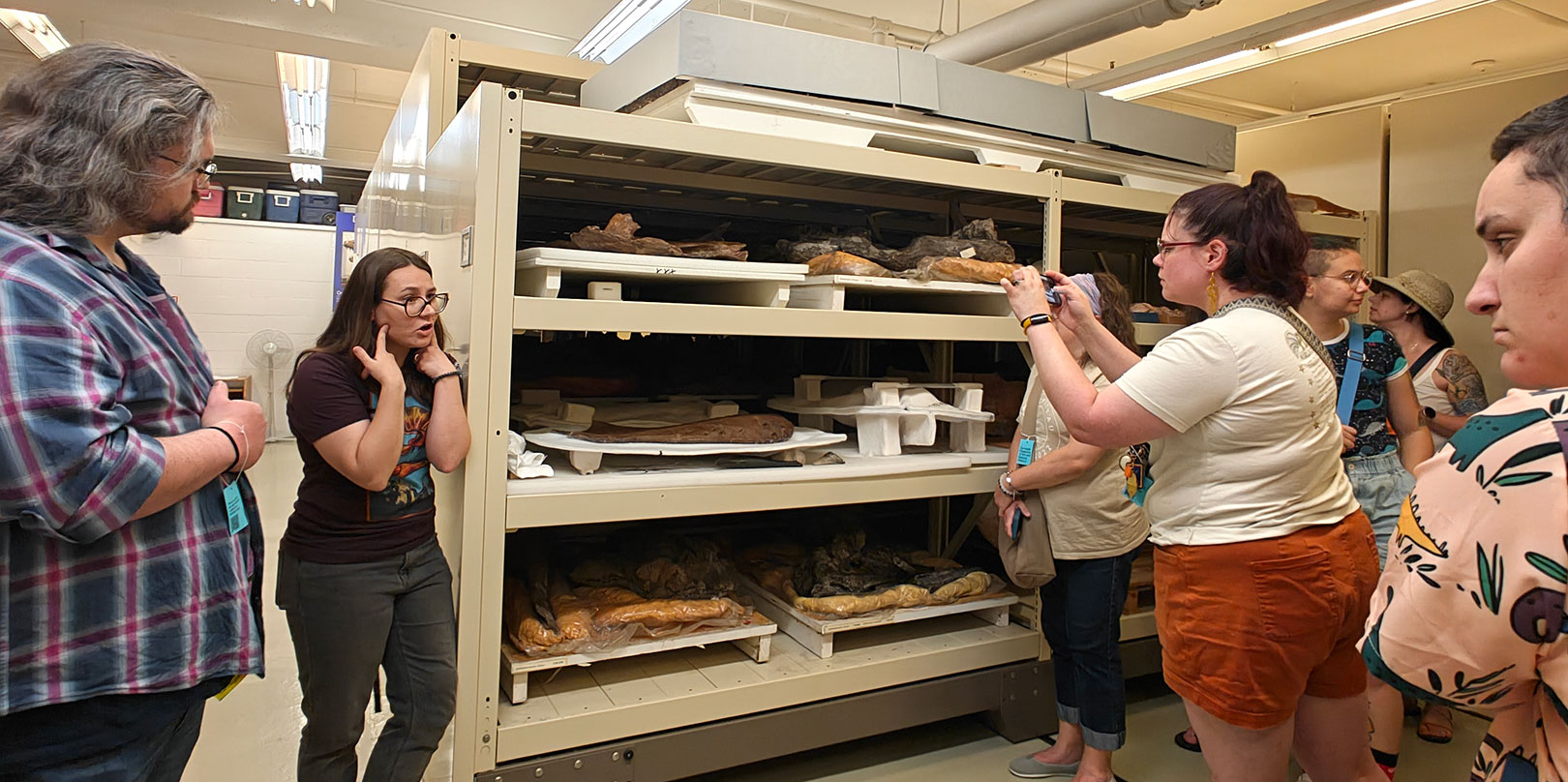 Visitors examine shelves of breads and pastries in a bakery display rack, while a woman photographs the baked goods in a busy, curious scene.