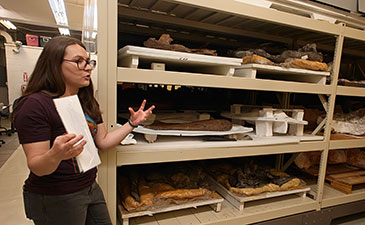 A woman gestures while explaining rows of bread and pastries on trays in a bakery storage rack, creating an informative, behind-the-scenes mood.