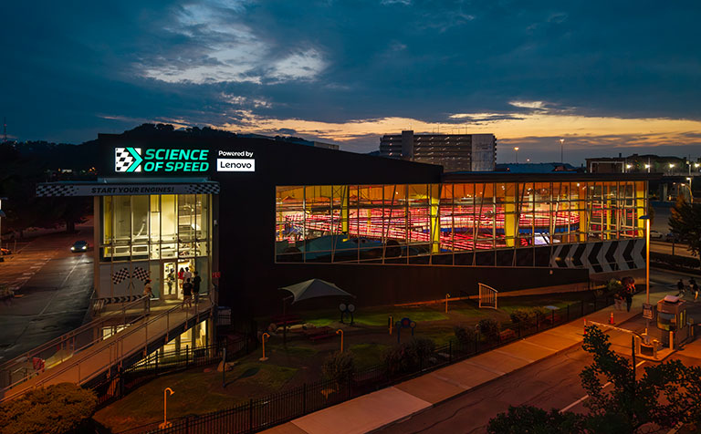 Dusk view of the Science of Speed exhibit, with glowing glass walls, visitors inside, walkways, and a dramatic cloudy sunset sky.