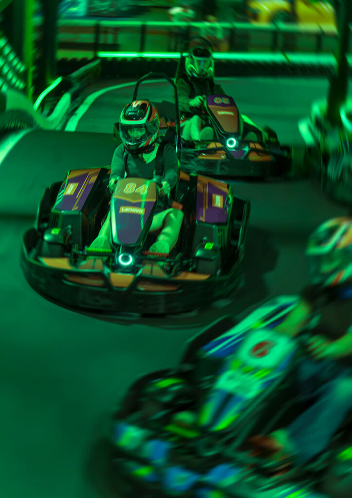 Two helmeted go-kart racers speed around a neon-green indoor track, with one kart sharply focused and another blurred in motion.