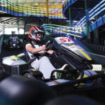Helmeted driver steers go-kart 51 around an indoor track, surrounded by tire barriers and colorful overhead lights in a fast-paced scene.
