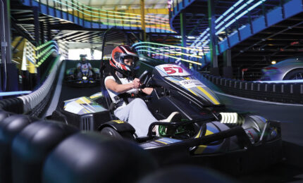 Helmeted driver steers go-kart 51 around an indoor track, surrounded by tire barriers and colorful overhead lights in a fast-paced scene.