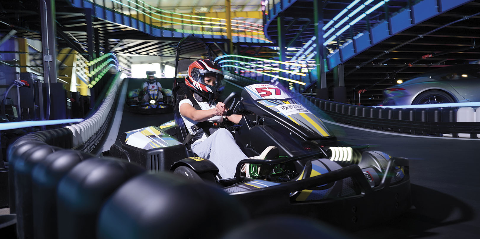 Helmeted driver steers go-kart 51 around an indoor track, surrounded by tire barriers and colorful overhead lights in a fast-paced scene.