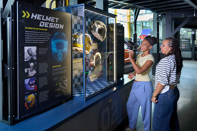 Two visitors with interest explore a motorcycle helmet design exhibit, viewing helmets in glass cases and an educational museum display.