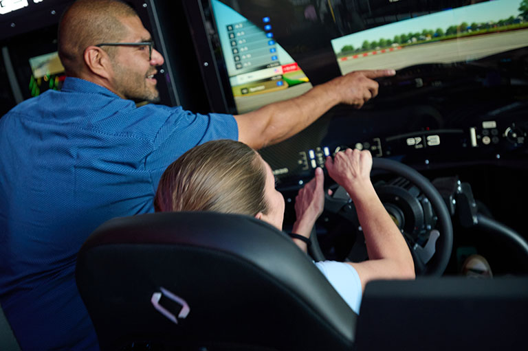 A smiling man points at a racing simulator screen while a woman grips the steering wheel, surrounded by dashboard controls in a dark cockpit.