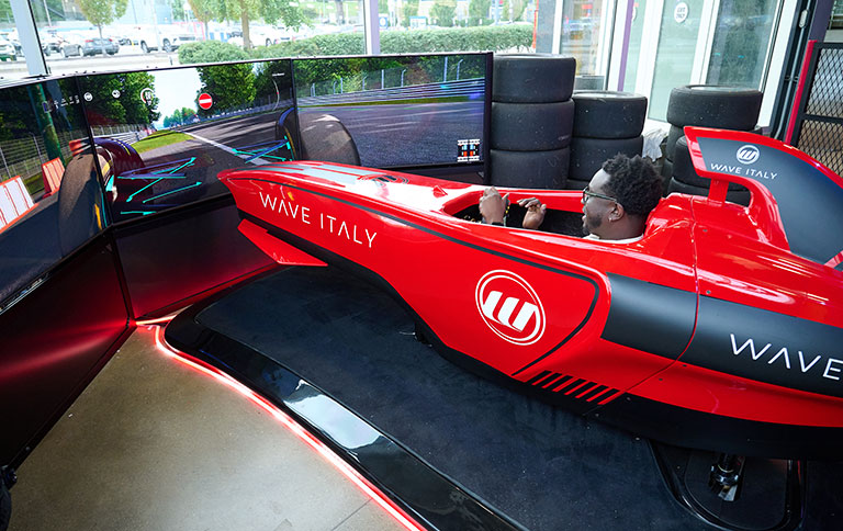 Person sitting in a red racing simulator car, holding the steering wheel and facing three large screens displaying a racetrack scene.