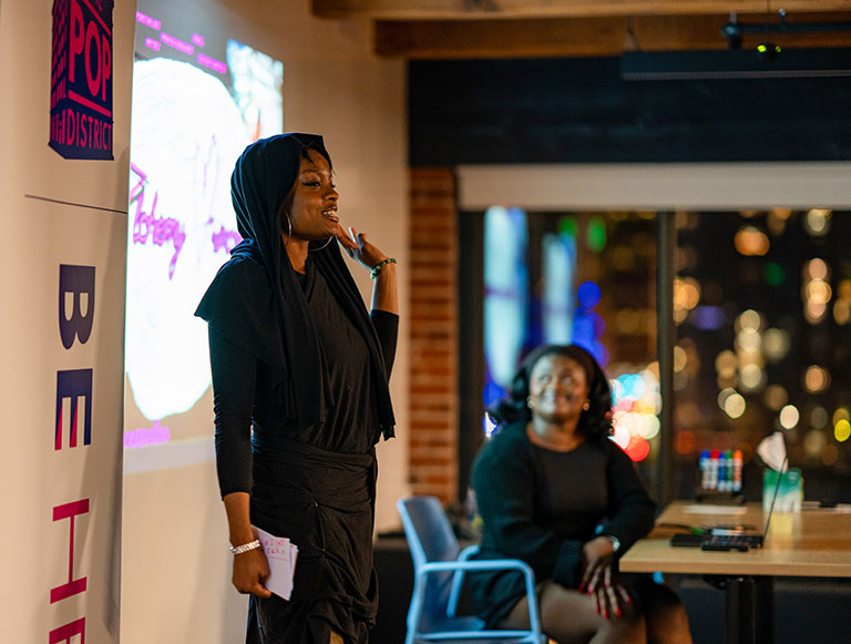 A woman in a black outfit and headscarf speaking at an indoor event, with another woman seated and city lights in the background.