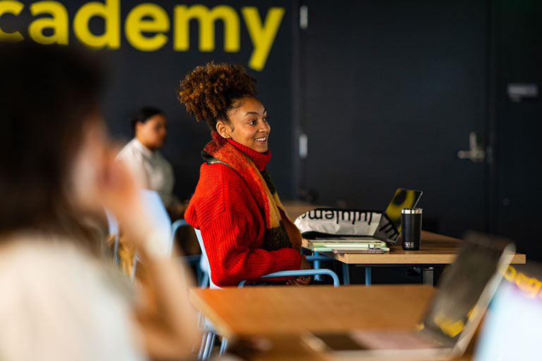 Young woman in a red sweater sits at a desk smiling, with laptops and a tote bag in a classroom setting with "academy" text on the wall.
