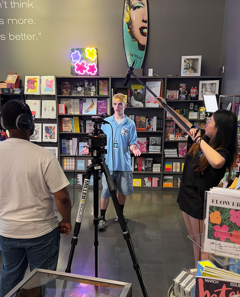 Three people film an interview inside a colorful art store with shelves of books and prints in the background.