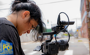Young person with dark hair operating a professional video camera on a city street during daytime.