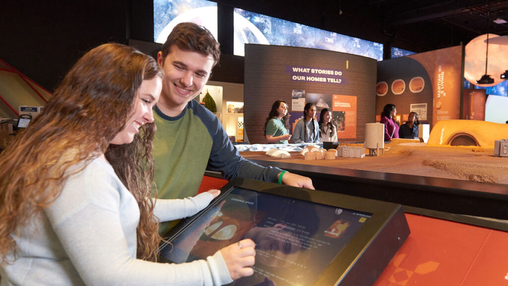 Two visitors interact with a touchscreen exhibit at a science museum, exploring displays about space and habitation on other planets.