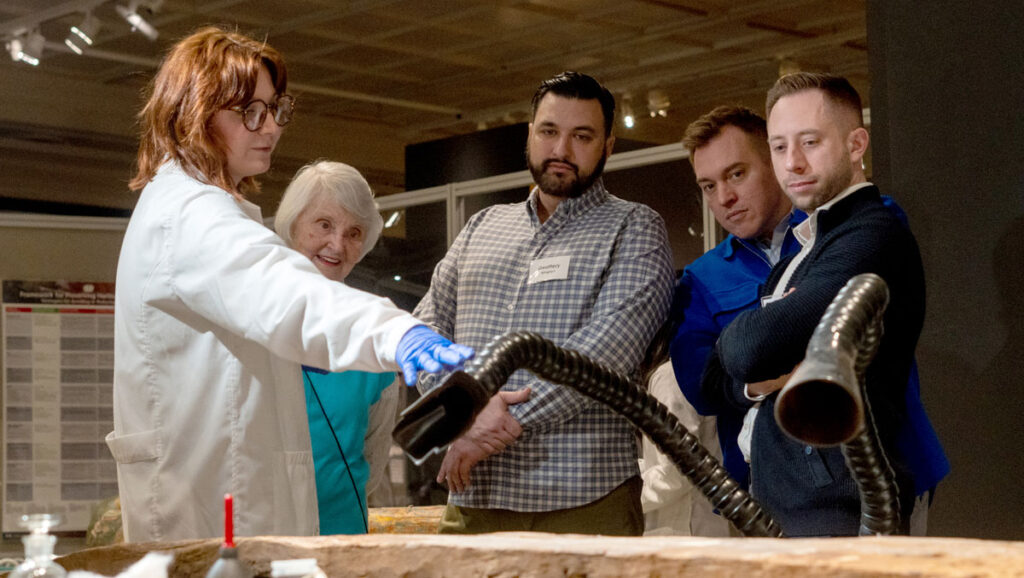 A scientist in a lab coat demonstrates alongside a wooden table, engaging a group of interested observers in an educational setting.