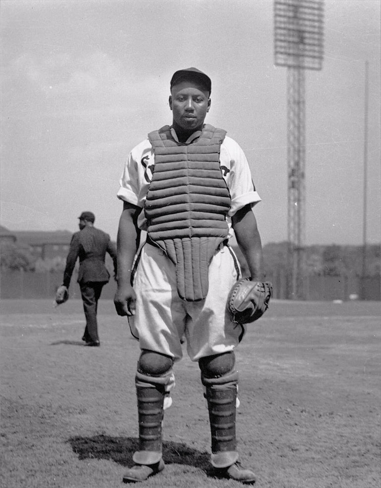 A black and white photo of an Black baseball player wearing a catchers vest, and a catcher's mitt. An umpire is walking in the background.