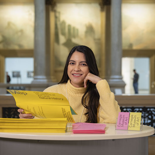 A person wearing a yellow sweater sits at a table, reading a large yellow brochure in a museum with art on display in the background.