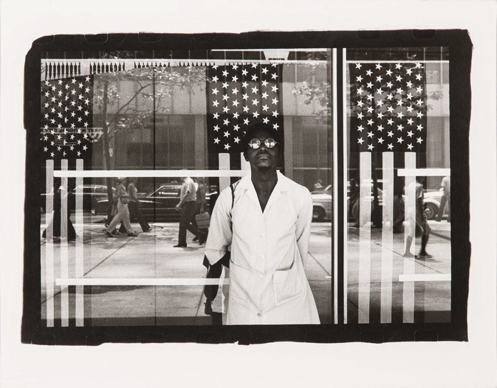 Black and white photo of a man standing in front of a window that's reflecting the american flag