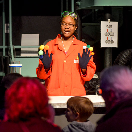 A person in an orange lab coat and gloves holds colorful balls, demonstrating a science experiment to an attentive audience.