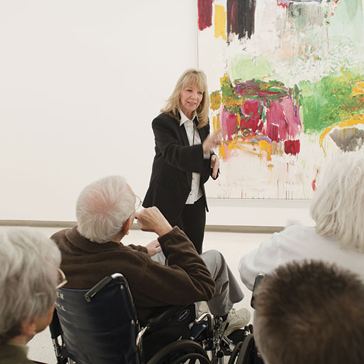 A woman gestures while presenting to an audience of elderly individuals in wheelchairs, with a colorful abstract painting in the background.
