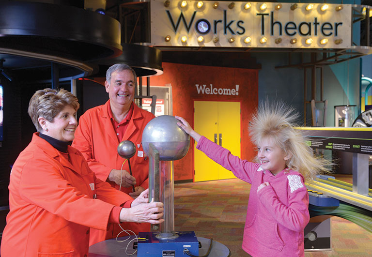 An adult couple standing next to a young girl with her hand on a van de Graff generator. The gilrs hair is standing on end from the static electricity.