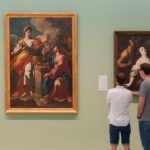 A art gallery image showing artwork against a green colored wall with two patrons looking at the artwork..