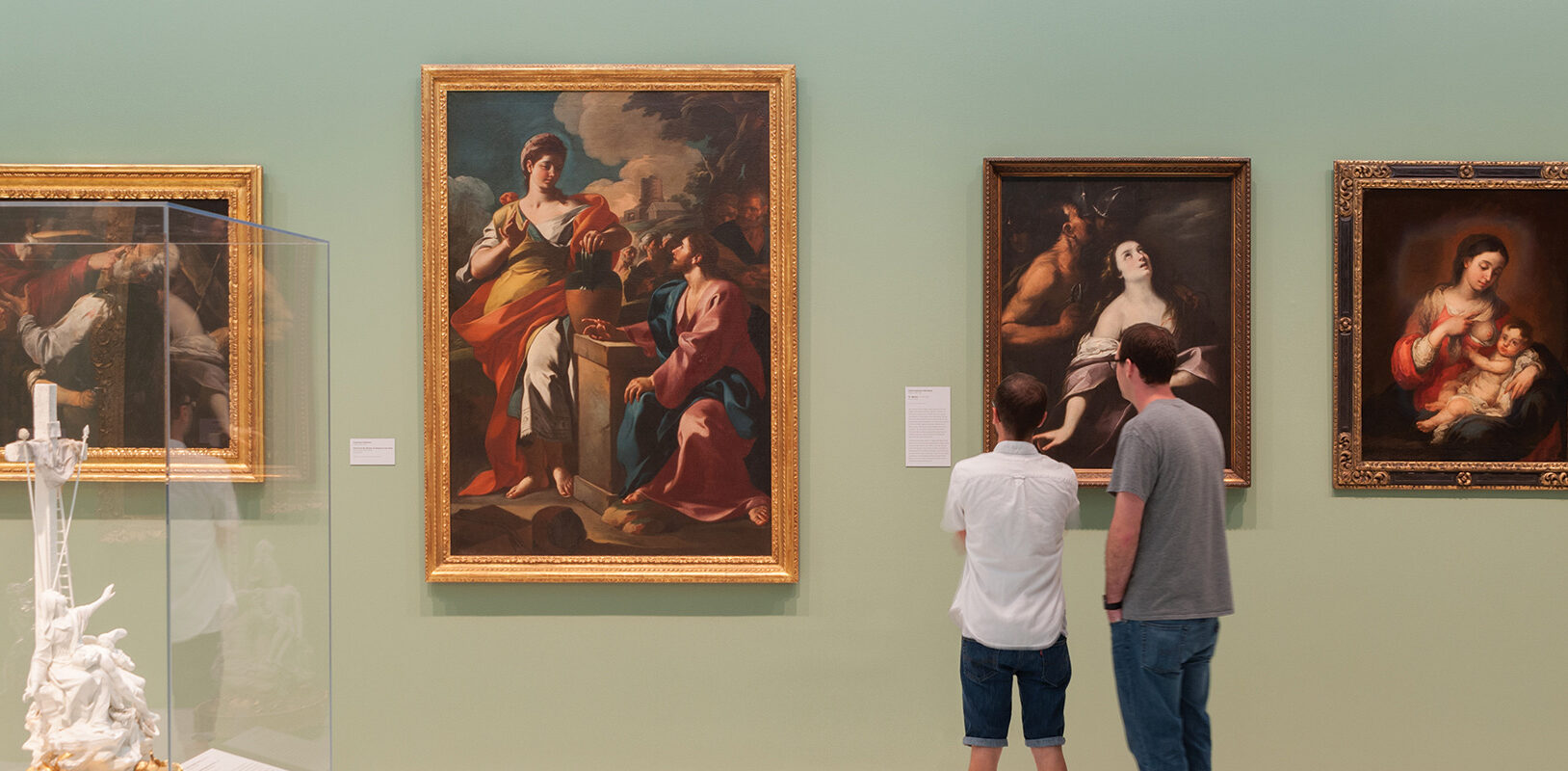 A art gallery image showing artwork against a green colored wall with two patrons looking at the artwork..