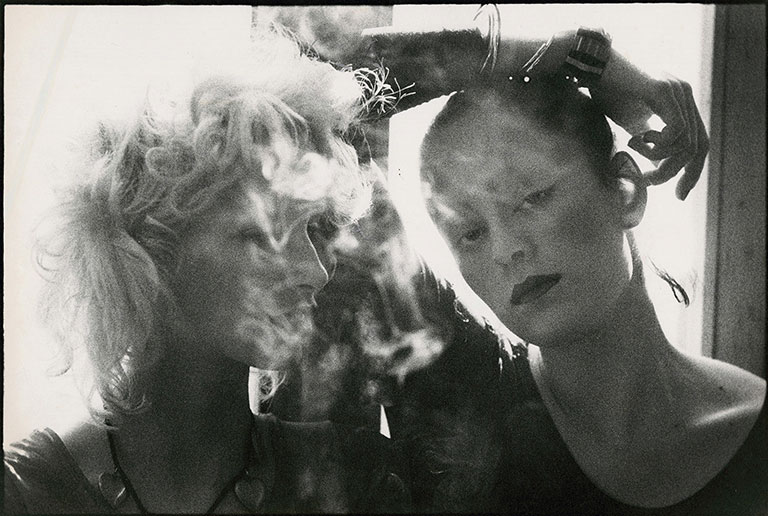 A black and white image of 2 women standing close together with cigarette smoke in the air around them