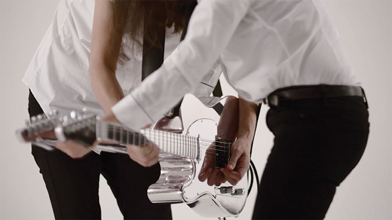A photo of 2 people facing each other playing the same guitar. THey are shown from the neck down and knees up