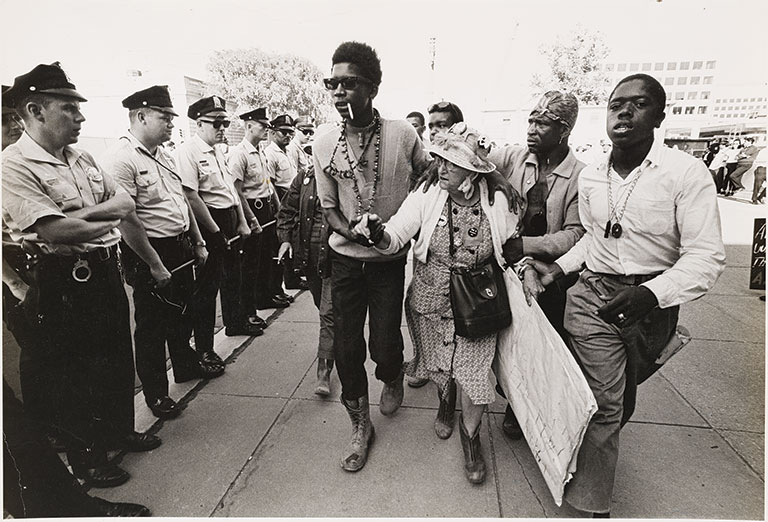 A group of men helping an elderly african american women past a line of policemen