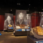 An installation view of an exhibition of war shields with large black and white photos of Native American women behind them