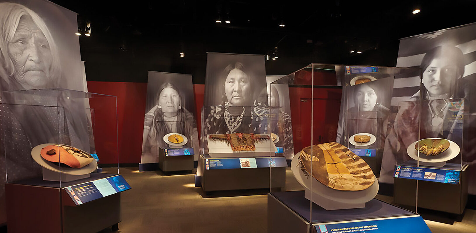 An installation view of an exhibition of war shields with large black and white photos of Native American women behind them