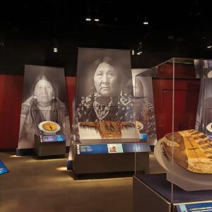 An installation view of an exhibition of war shields with large black and white photos of Native American women behind them