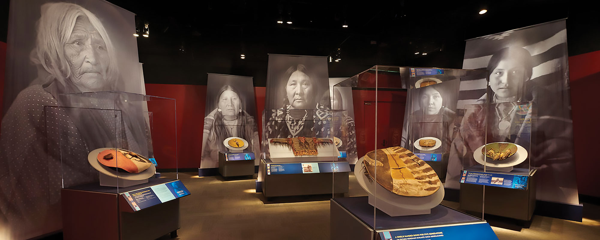 An installation view of an exhibition of war shields with large black and white photos of Native American women behind them