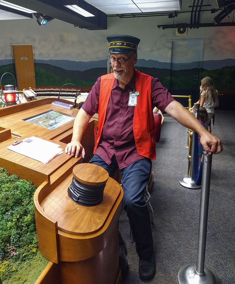 A man wearing a conductor's hat sitting at the controls of the miniature railroad.