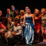 A diverse group of people in creative, colorful clothing pose together on stage, smiling. The mood is celebratory and theatrical, with dramatic lighting.
