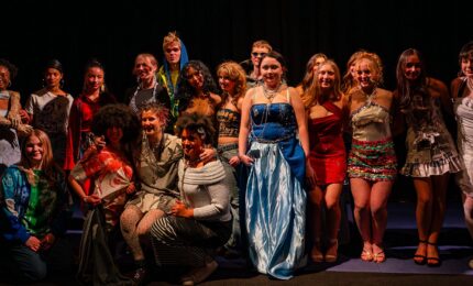 A diverse group of people in creative, colorful clothing pose together on stage, smiling. The mood is celebratory and theatrical, with dramatic lighting.