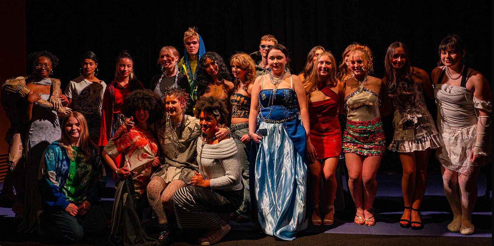 A diverse group of people in creative, colorful clothing pose together on stage, smiling. The mood is celebratory and theatrical, with dramatic lighting.