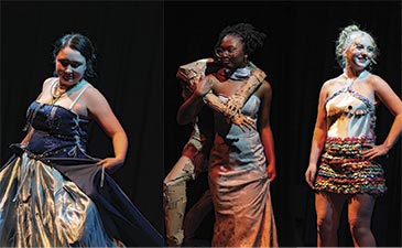 Three people on stage wearing creative costumes. One in a blue gown, another in a textured dress with a cardboard arm piece, and the third in a colorful skirt.