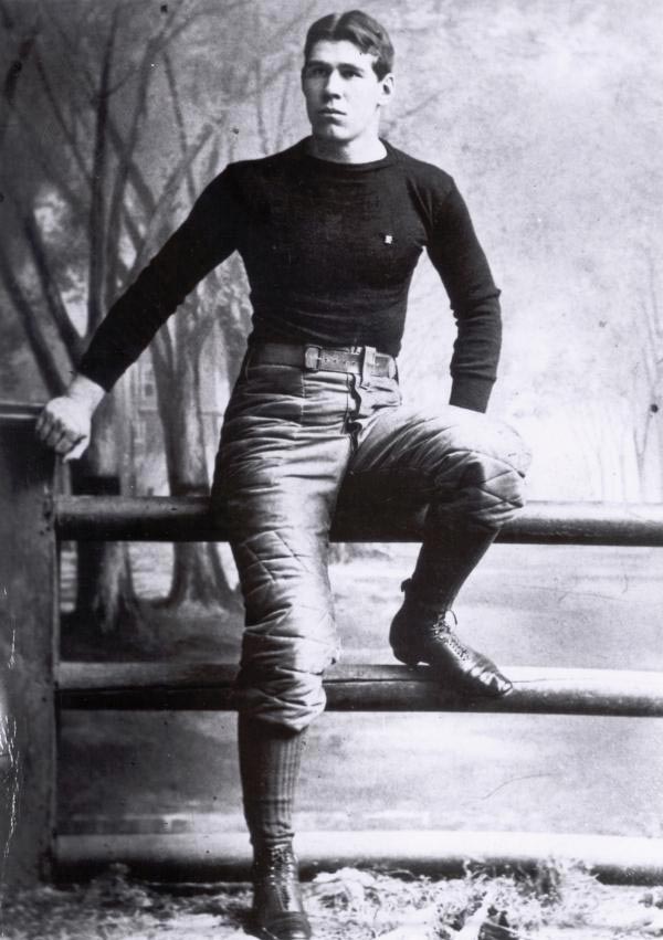A young man in vintage football attire leans on a wooden fence in a forest setting. He exudes a confident, serious expression in this black-and-white photo.