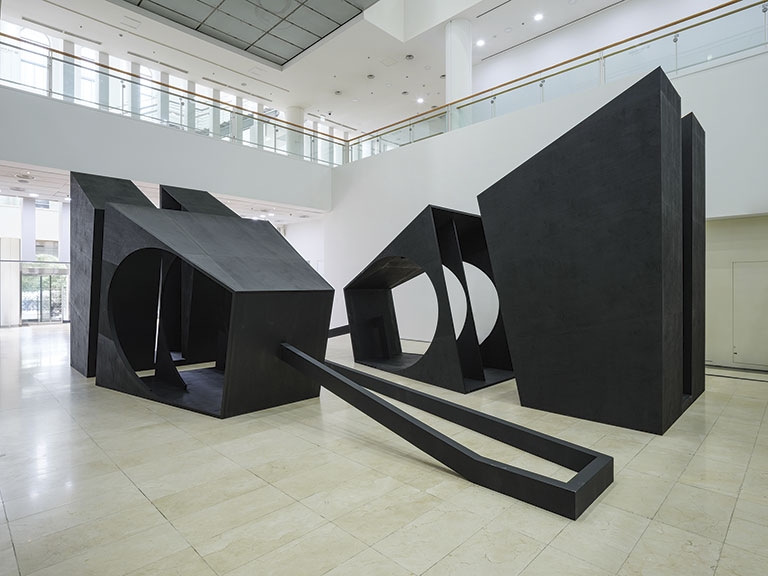 Modern art installation in a spacious gallery, featuring large geometric black structures with circular cutouts on a polished floor. The space feels minimalistic.