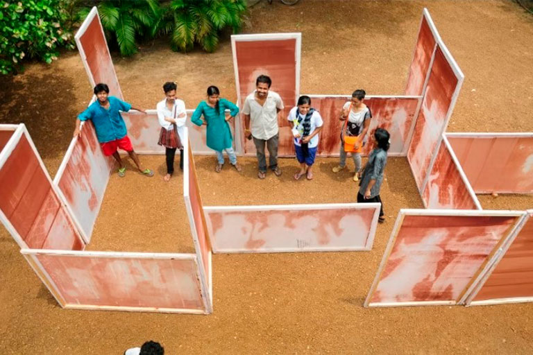 A group of seven people stands smiling within a maze-like structure made of red, patched panels on a dirt ground, conveying a playful, collaborative atmosphere.