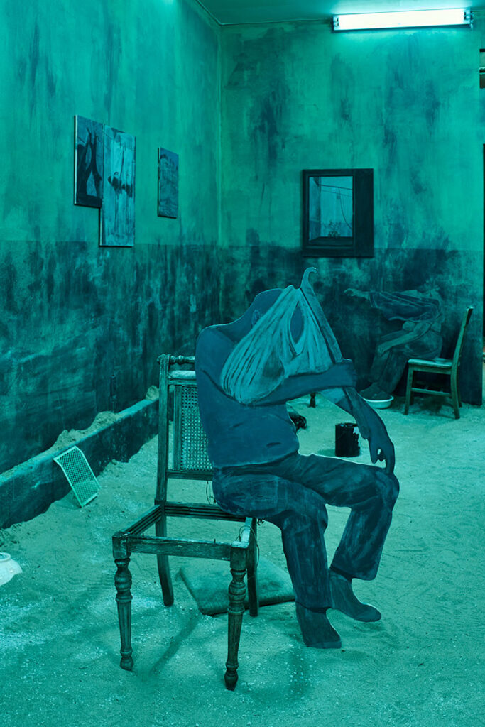 Dimly lit blue-green room with paintings on walls. A painted figure slumps on a chair, creating a surreal, melancholic atmosphere.
