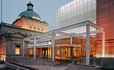 Grand building with a domed roof and modern glass extension houses a children's museum. Warm lighting creates an inviting atmosphere at dusk.